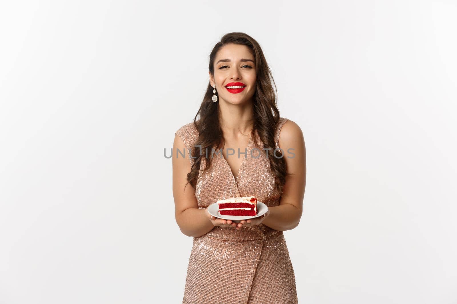 Party and celebration concept. Attractive woman in elegant dress holding delicious cake and smiling with temptation, standing over white background.
