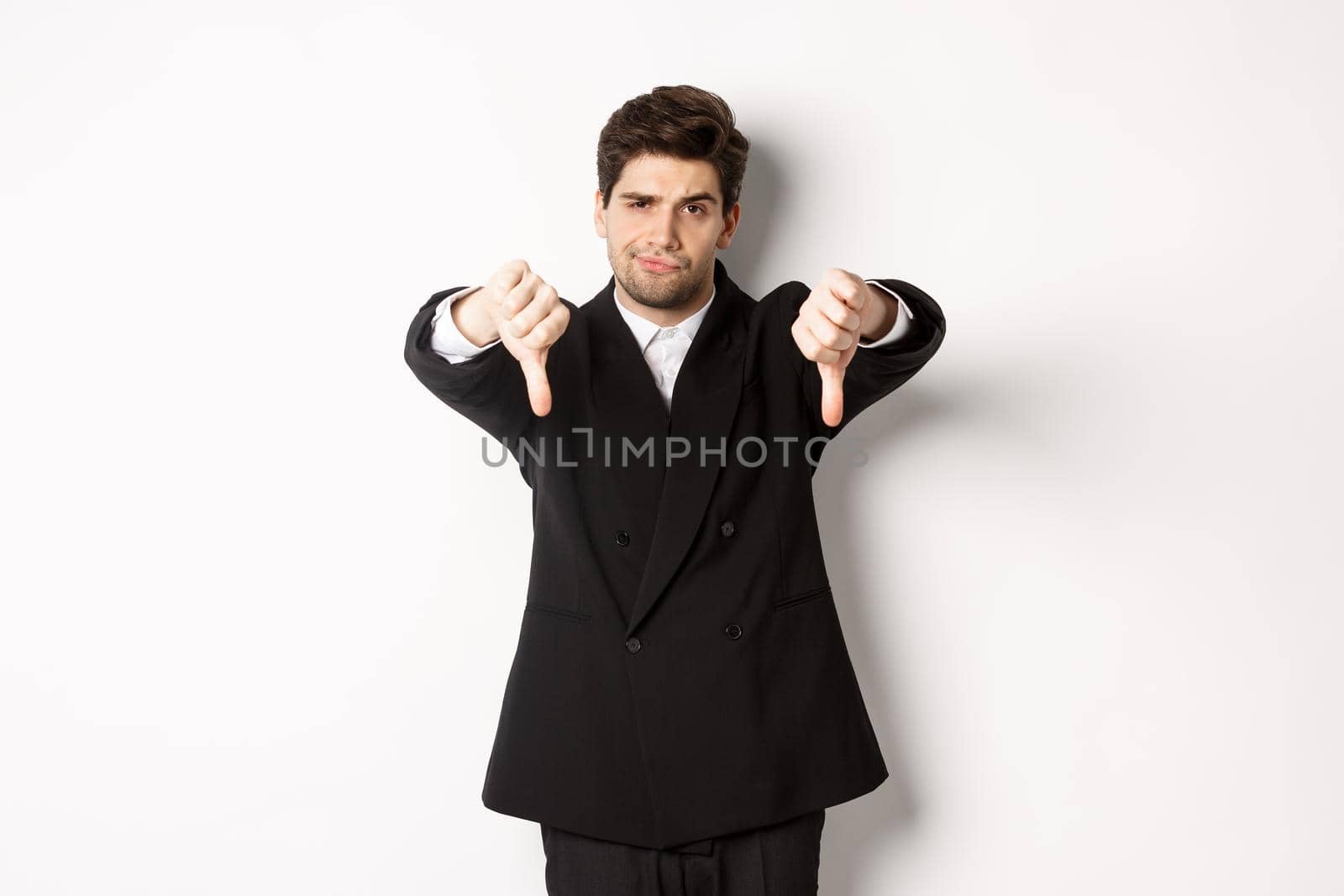 Portrait of skeptical and disappointed man in black suit, frowning upset, showing thumbs-down, dislike something bad, standing over white background.