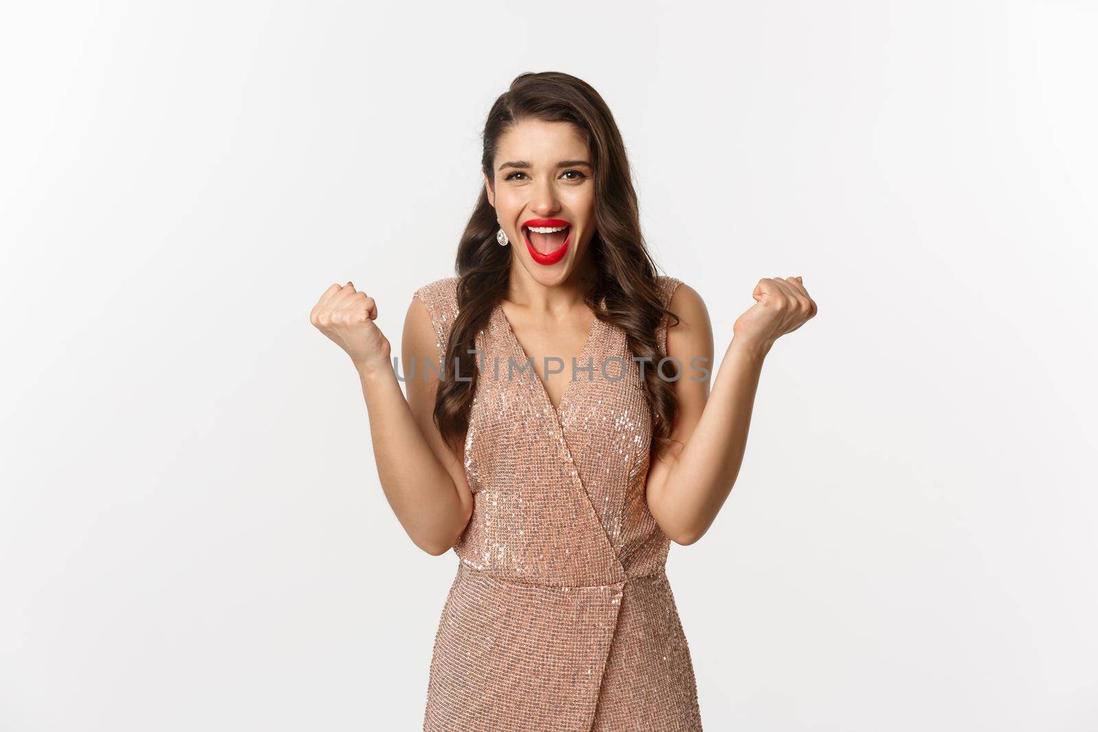 Celebration and party concept. Elegant young woman winning and feeling satisfied, wearing glamour dress, making fist pump and say yes, triumphing over white background.