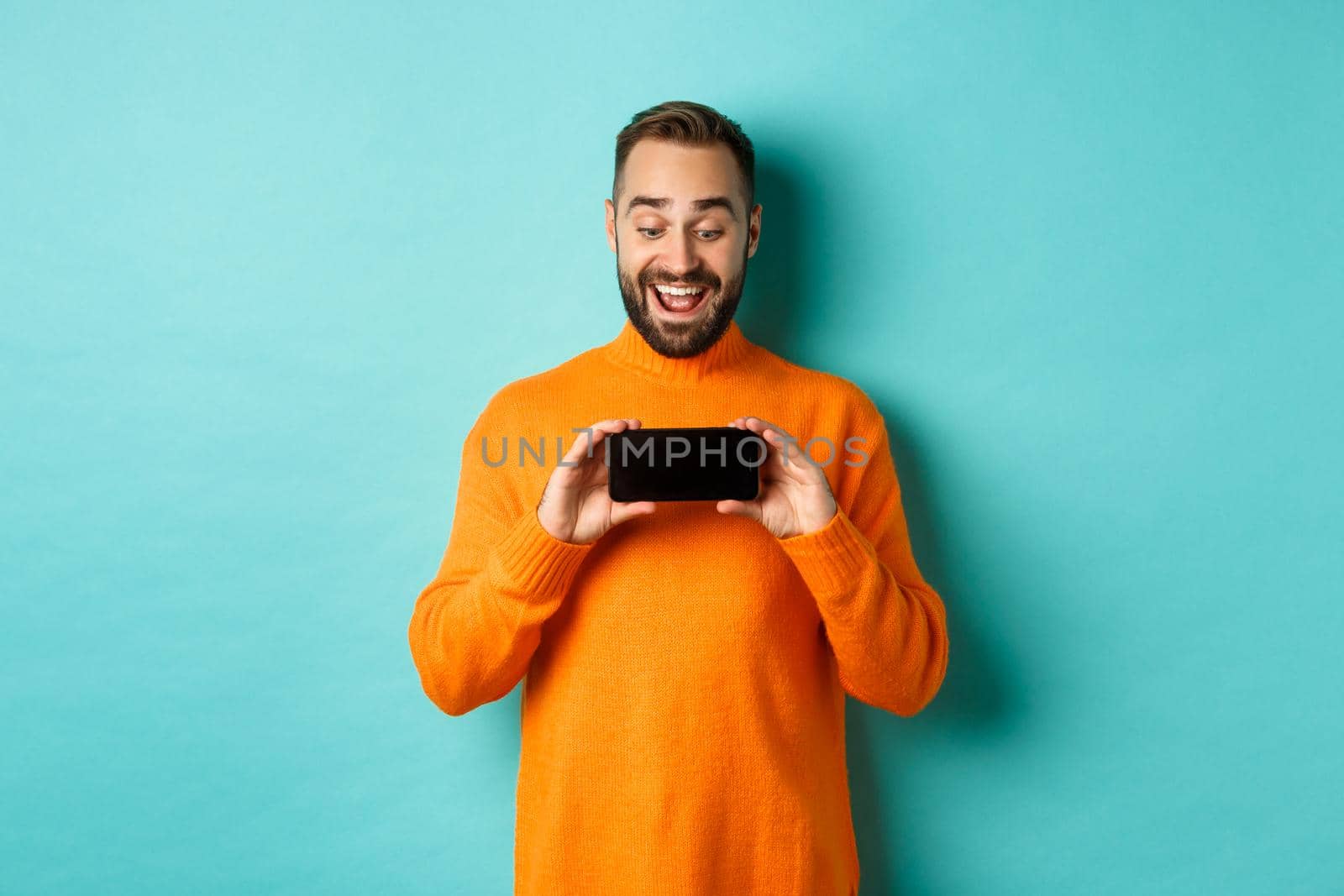Excited guy demonstrating online store, showing mobile screen and looking amazed, standing over light blue background.