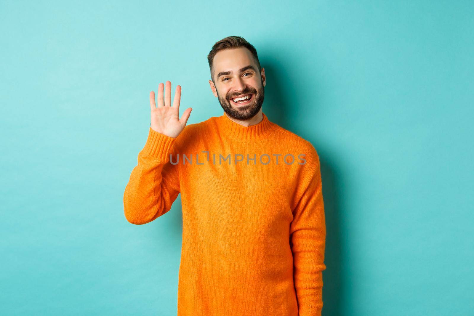 Photo of friendly young man saying hello, smiling and waiving hand, greeting you, standing in orange sweater over light blue background.