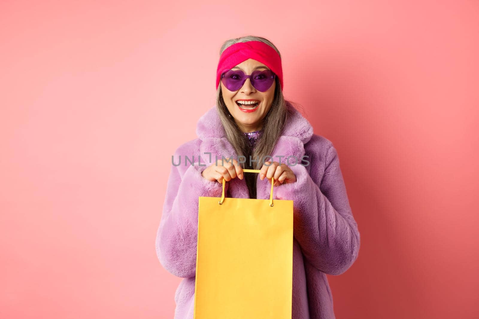 Fashionable asian senior woman going shopping in sunglasses and trendy winter clothes, holding paper bag from store and smiling happy at camera, pink background by Benzoix