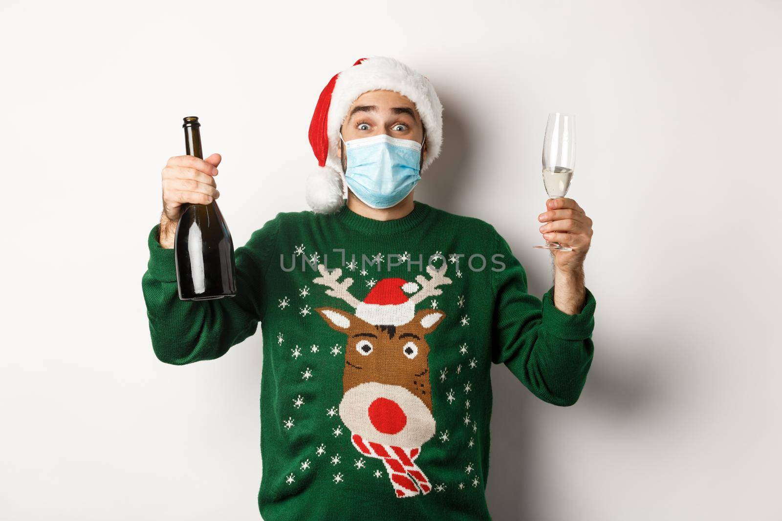 Concept of covid-19 and Christmas holidays. Happy man in face mask and Santa hat celebrating New Year with champagne, standing over white background.
