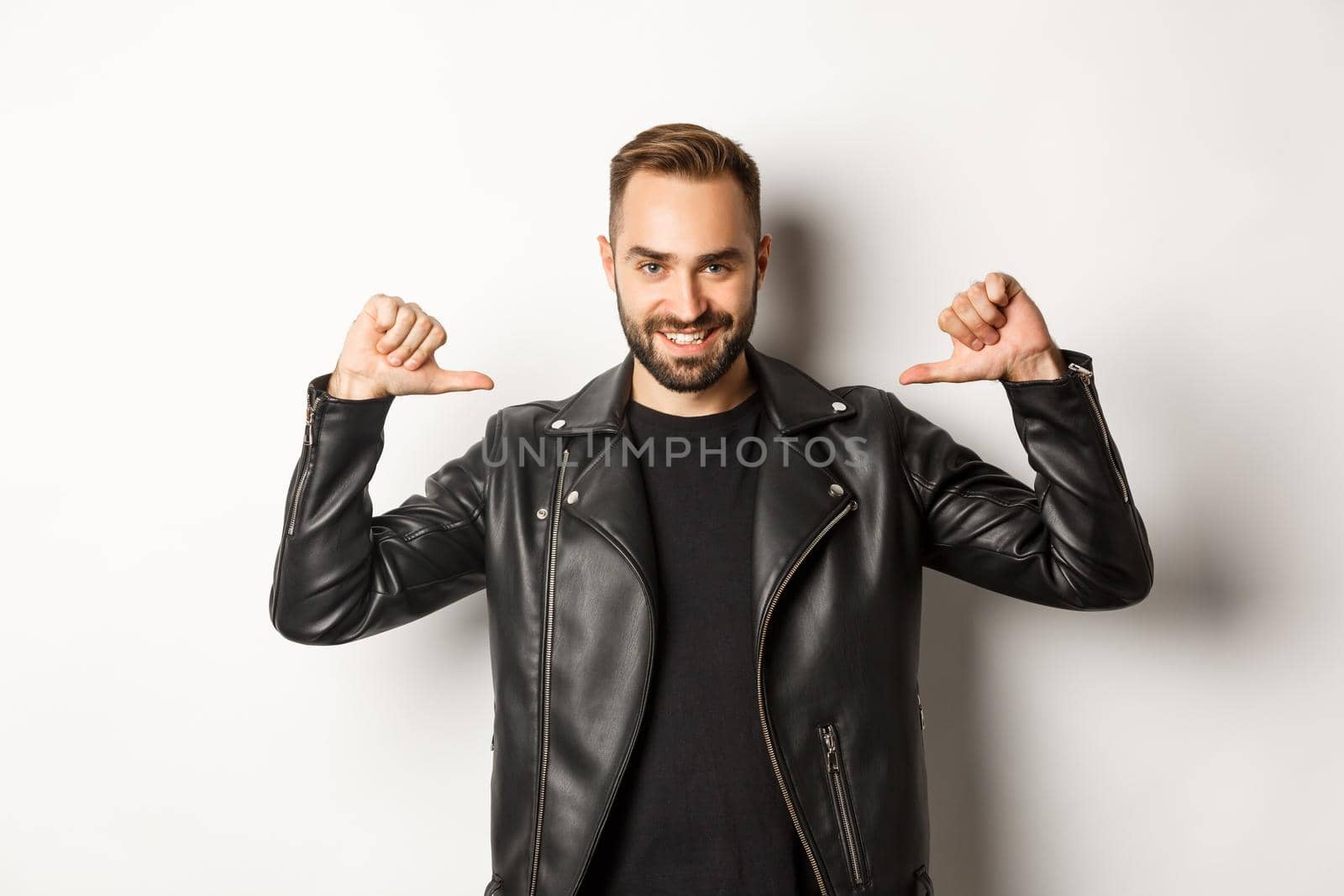 Confident handsome man wearing black leather jacket, pointing at himself and smiling self-assured, standing against white background.