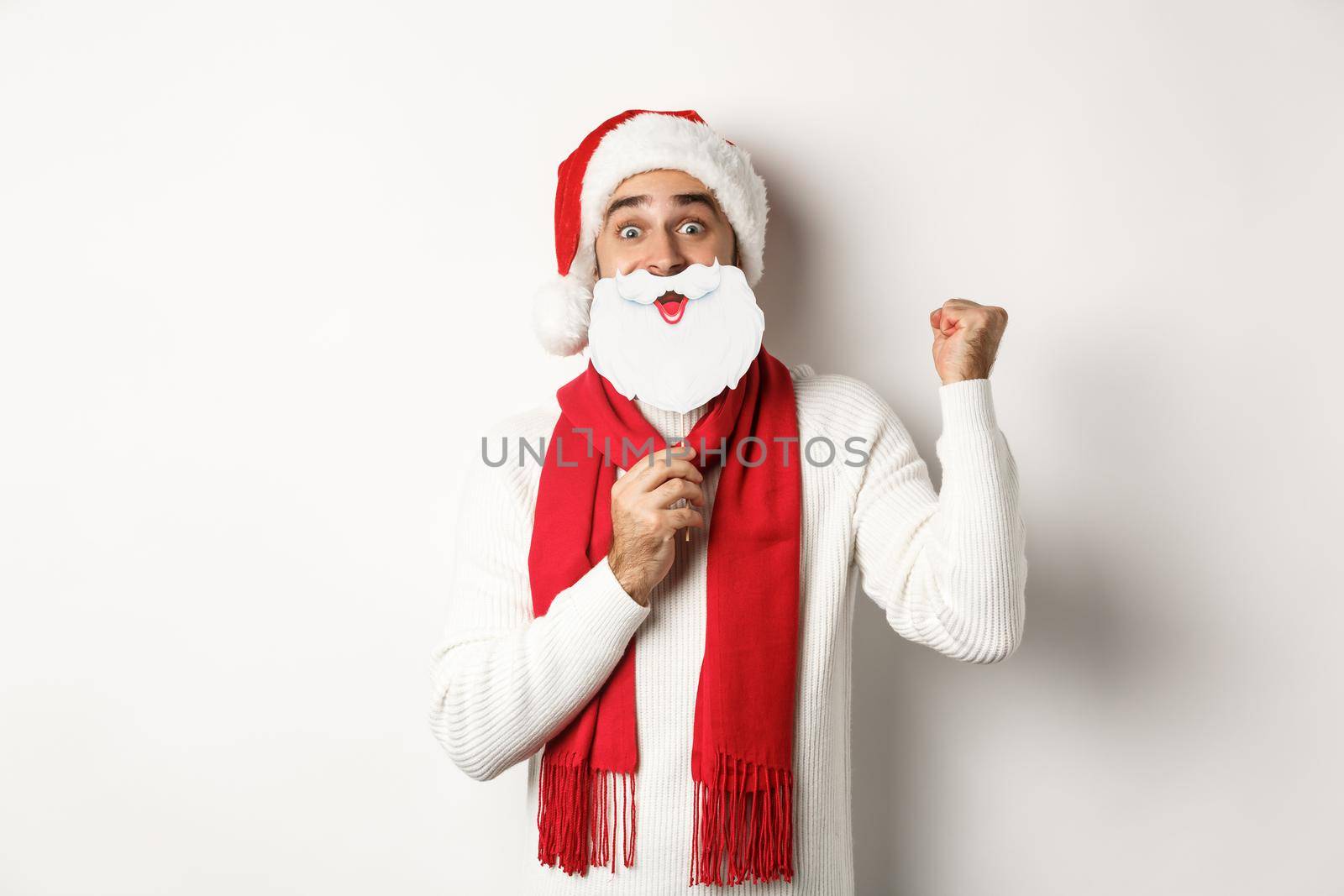 Christmas party and celebration concept. Excited young man enjoying New Year, looking funny in Santa hat with white beard mask, making fist pump and rejoicing.