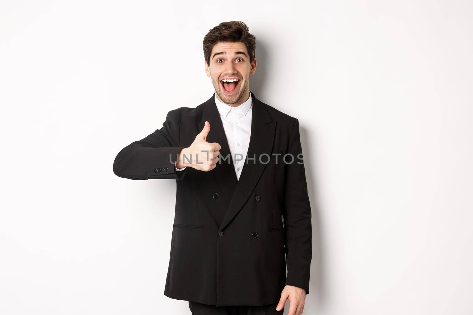 Portrait of amazed and impressed handsome man in black suit, showing thumb-up, praise awesome product, liking something good, standing against white background by Benzoix