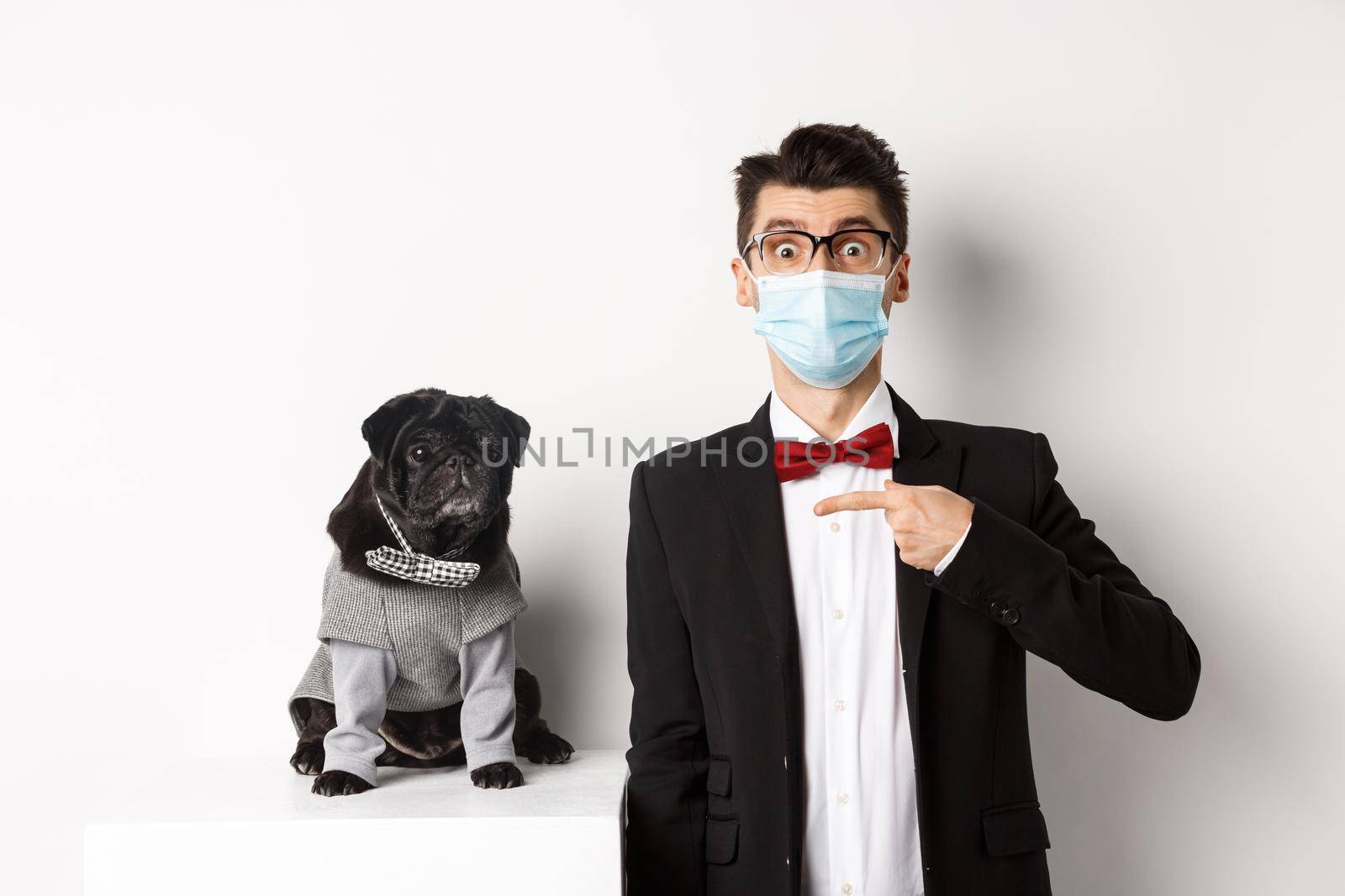 Coronavirus, pets and celebration concept. Amazed young man in face mask and suit pointing at cute black dog sitting near owner in party outfit, white background.