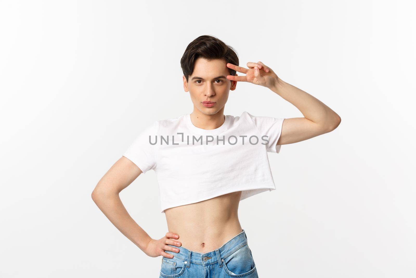 Beautiful androgynous man in crop top showing peace sign and smiling, standing over white background.