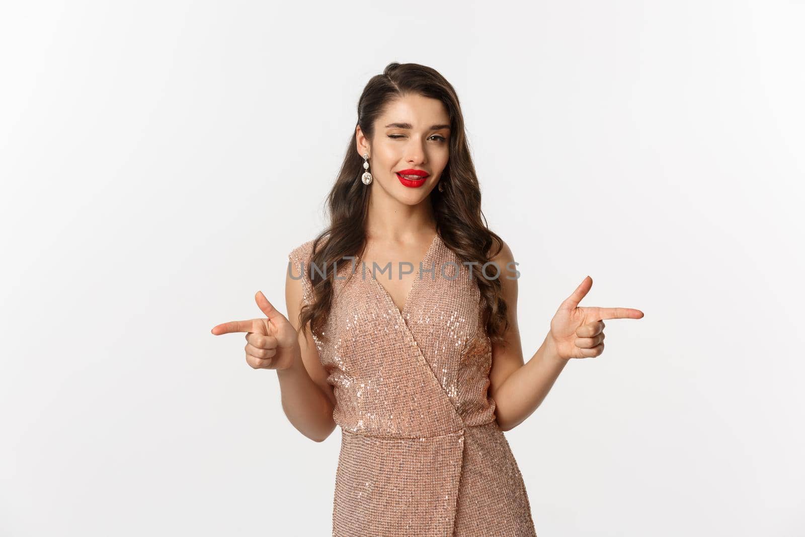 Concept of celebration, holidays and party. Beautiful and flirty woman with red lips, wearing glamour dress and winking, pointing fingers at camera, standing over white background by Benzoix