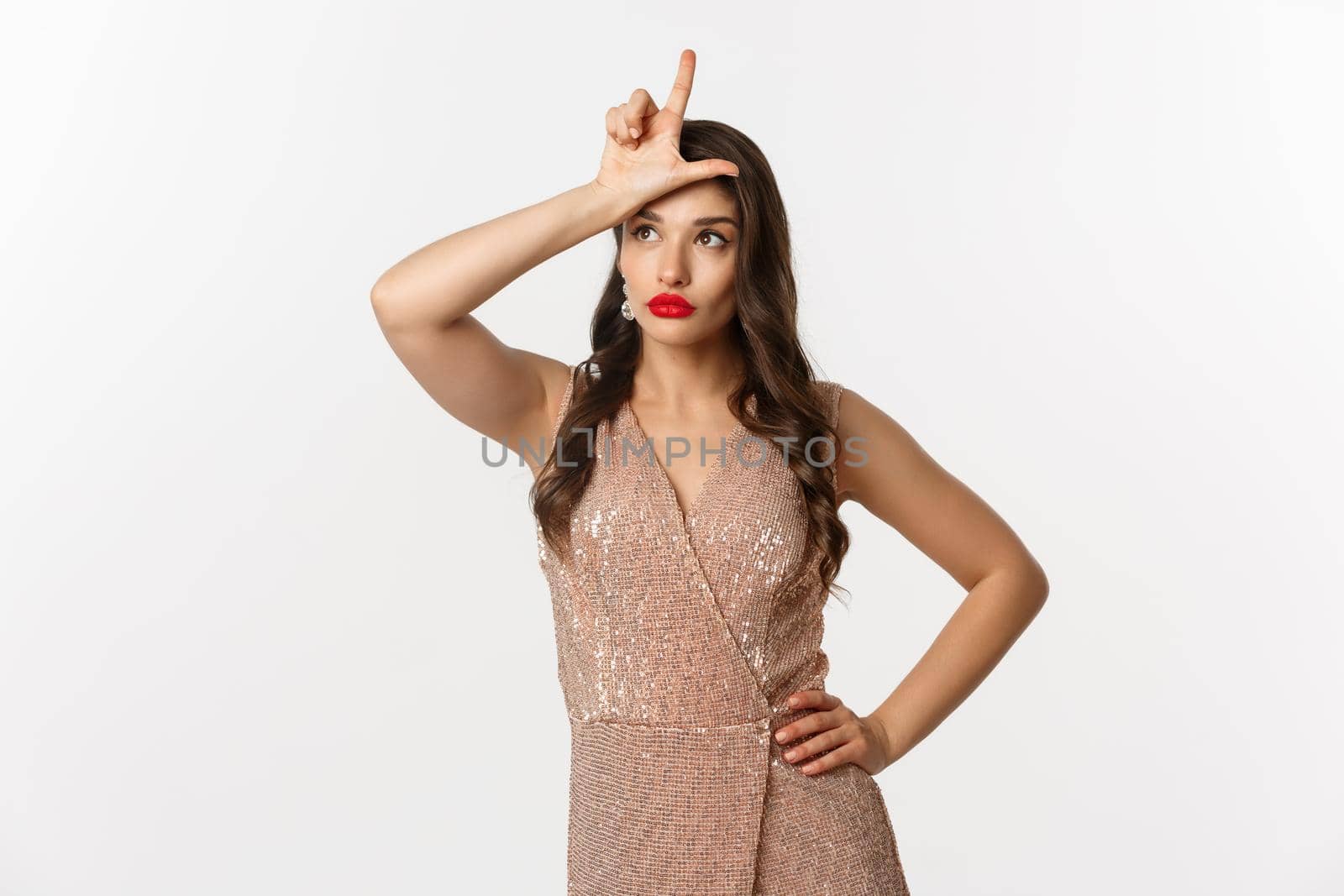 Celebration and party concept. Attractive young woman with red lips, glamour dress, showing loser sign on forehead and looking away ignorant, standing over white background.