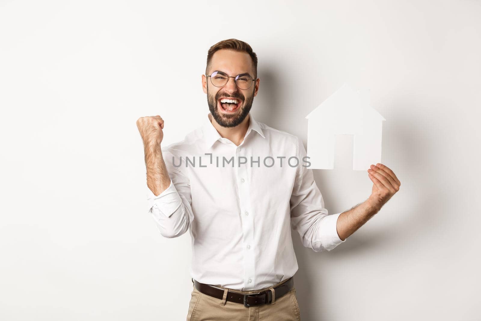Real estate. Satisfied man rejoicing of founding perfect home apartment, holding paper house model, standing over white background.