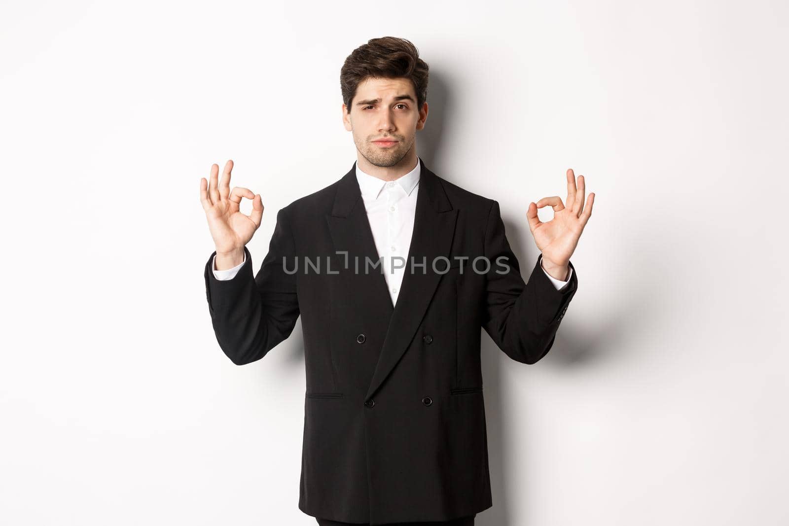 Concept of new year party, celebration and lifestyle. Portrait of confident good-looking man in black suit, showing okay sign and approve something, standing over white background.