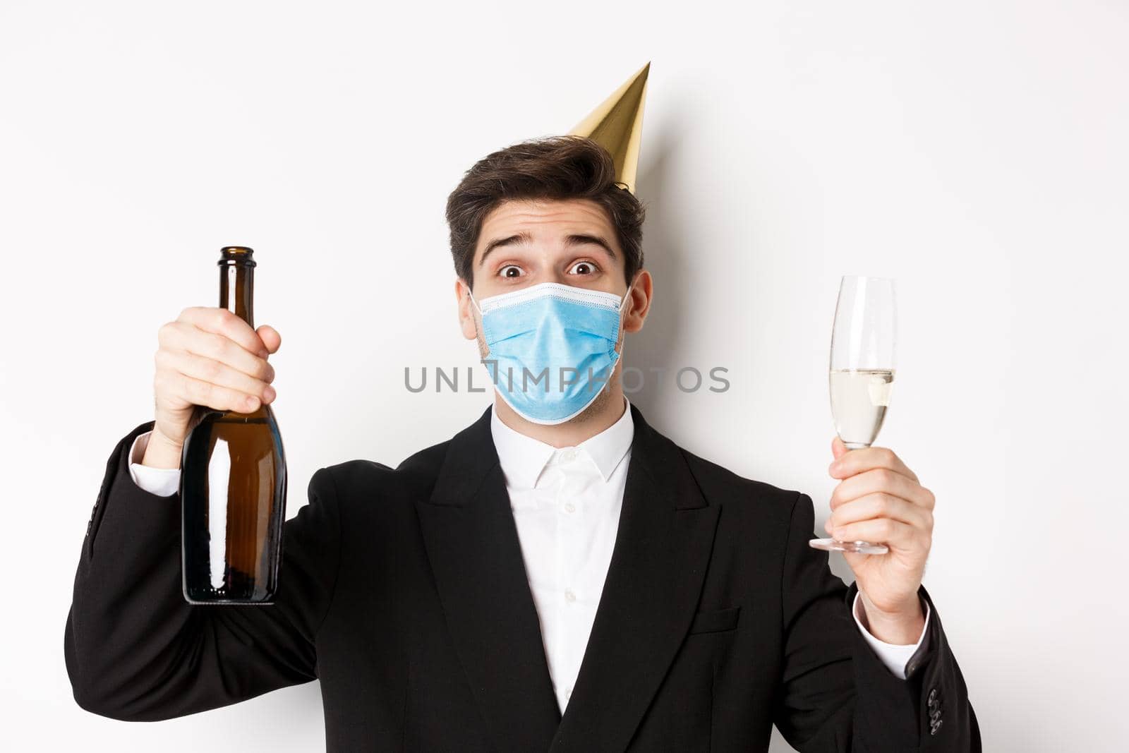 Concept of party during covid-19. Close-up of handsome man in suit, funny hat and medical mask, holding bottle of champagne, celebrating new year on social distancing.