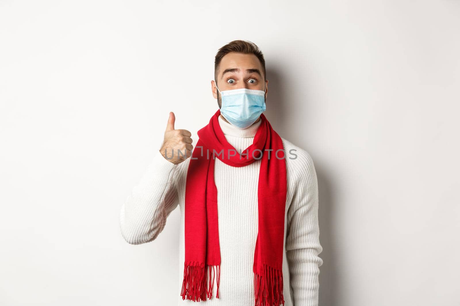 Covid-19, lockdown and quarantine concept. Amazed and satisfied man buyer in medical mask showing thumb-up, looking excited, like something good, white background.
