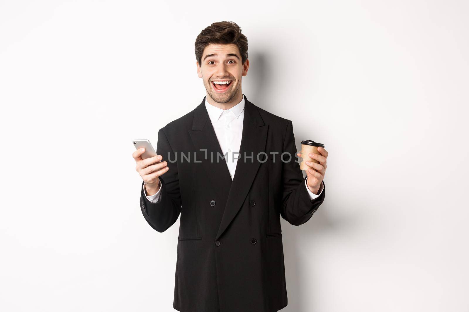Portrait of happy good-looking man in suit, holding cup of coffee and smartphone, achieve app goal, standing over white background.