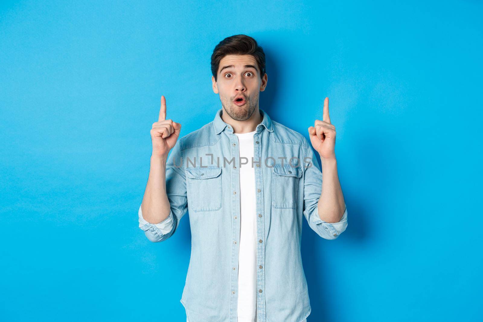 Portrait of amazed handsome man pointing fingers up, showing logo on blue background.