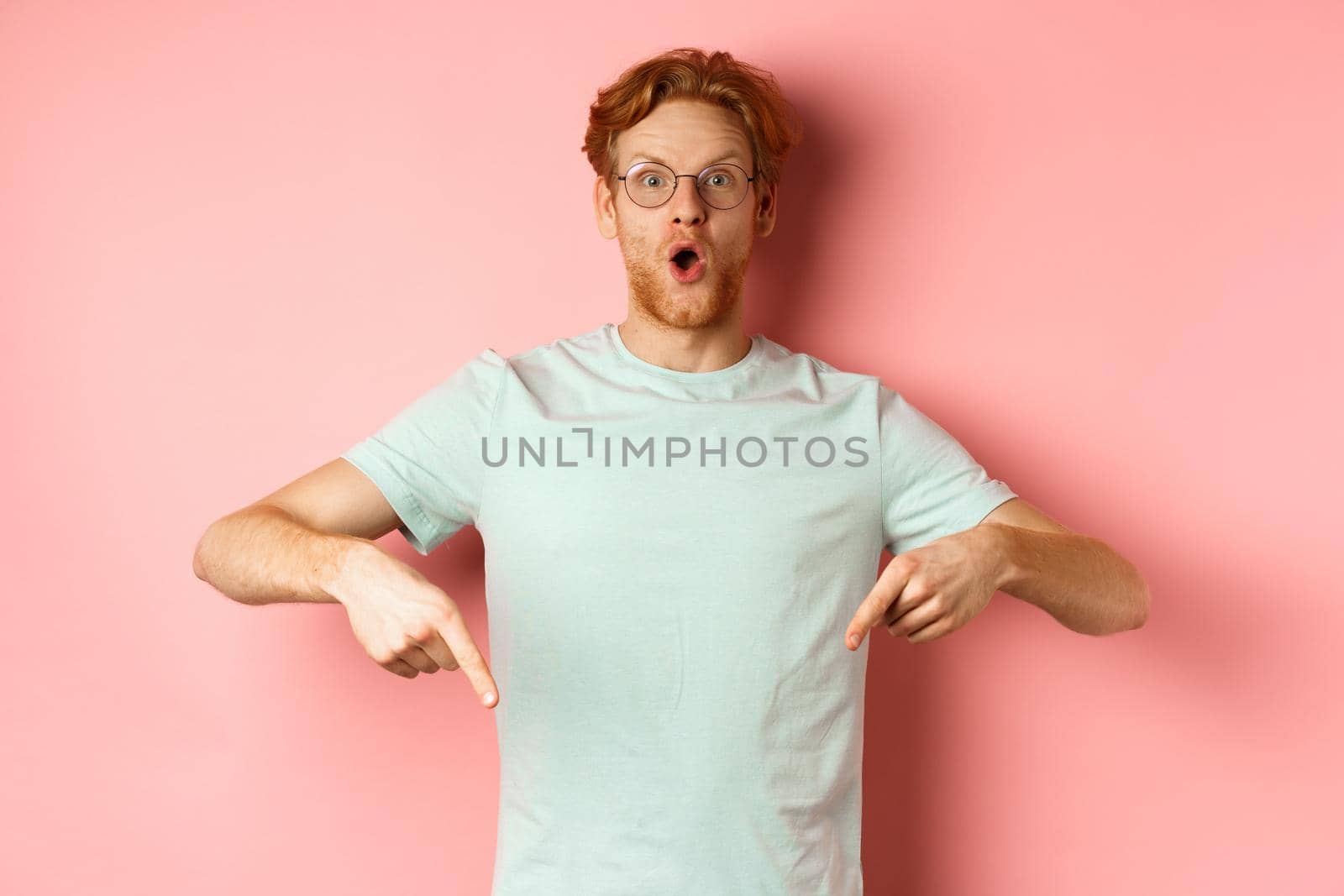 Image of amazed young man with red hair and beard, wearing glasses and t-shirt, pointing fingers down and staring excited at camera, pink background.