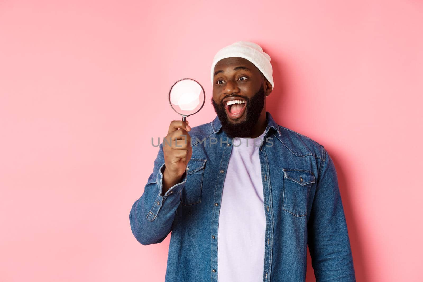 Happy african-american man looking through magnifying glass, smiling amazed, standing against pink background. Copy space