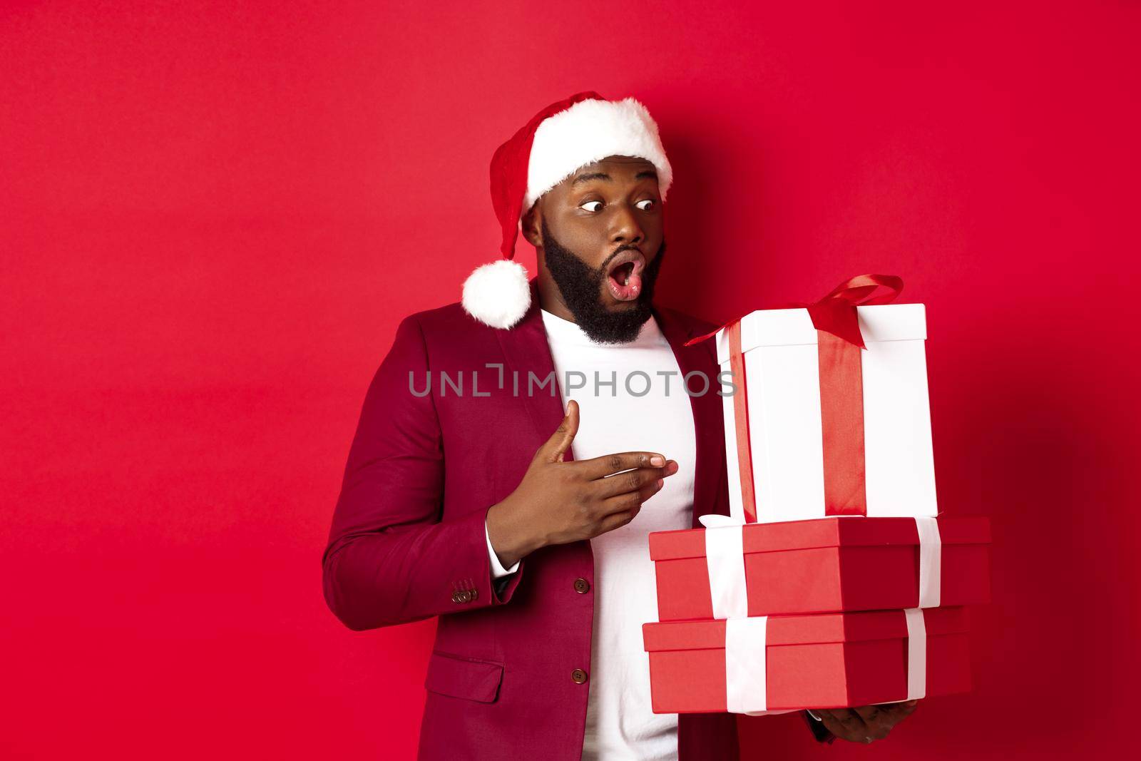 Christmas, New Year and shopping concept. Surprised Black man looking at xmas presents with amazement, wearing santa hat, standing with gifts against red background.