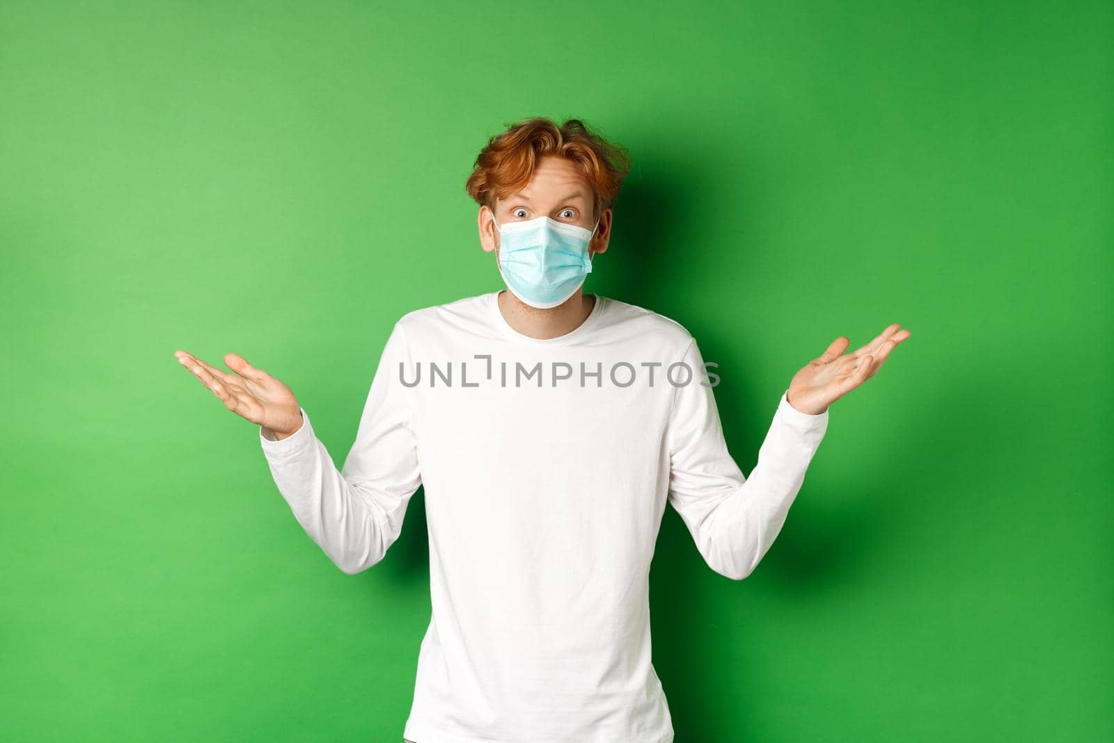 Covid-19, social distancing and lifestyle concept. Surprised redhead man in face mask spread hands sideways, staring at camera confused, standing over green background.