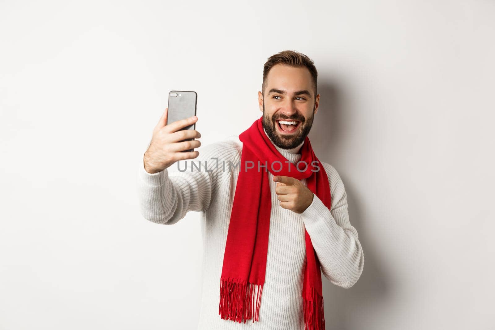 Handsome guy wishing merry christmas on video call, waving hand at mobile phone and smiling, standing in sweater with red scarf, white background.