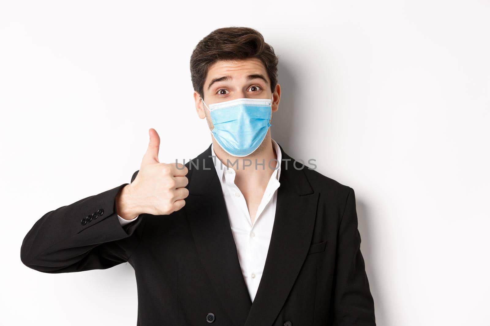 Concept of covid-19, business and social distancing. Close-up of happy businessman in black suit and medical mask, showing thumbs-up, making a compliment, white background.