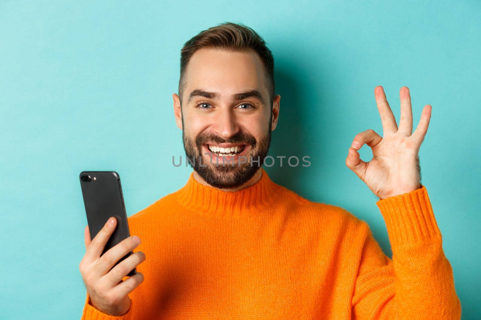 Close-up of bearded man holding smartphone and showing okay sign, recommending mobile app, standing satisfied over turquoise background.