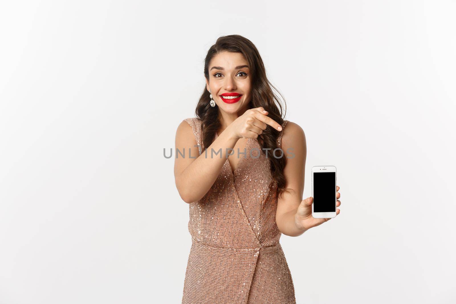 Online shopping. Excited young woman with red lips, elegant dress, pointing finger at mobile screen, showing something, standing over white background by Benzoix