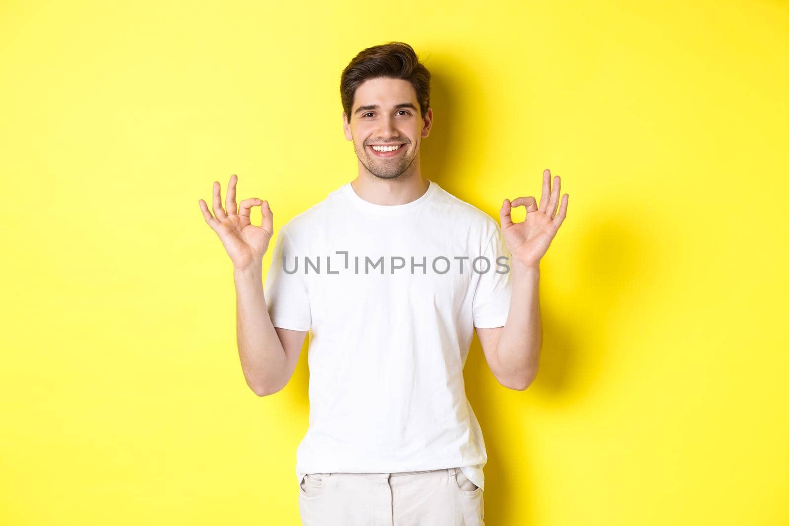 Relaxed guy smiling, showing okay signs, approve or agree, standing against yellow background.