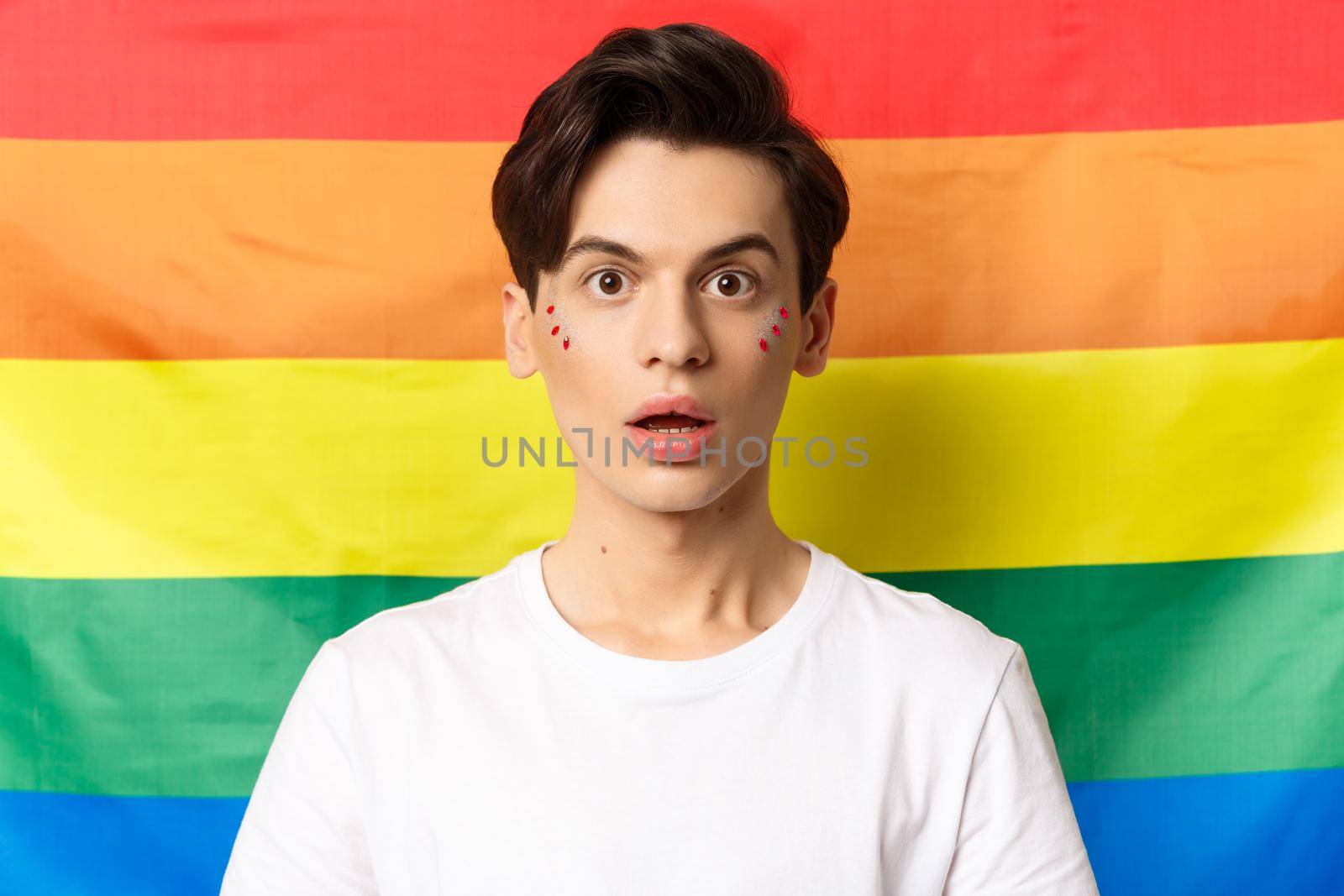 Close-up of surprised queer person with glitter on face, looking amazed at camera, standing over lgbtq rainbow flag.