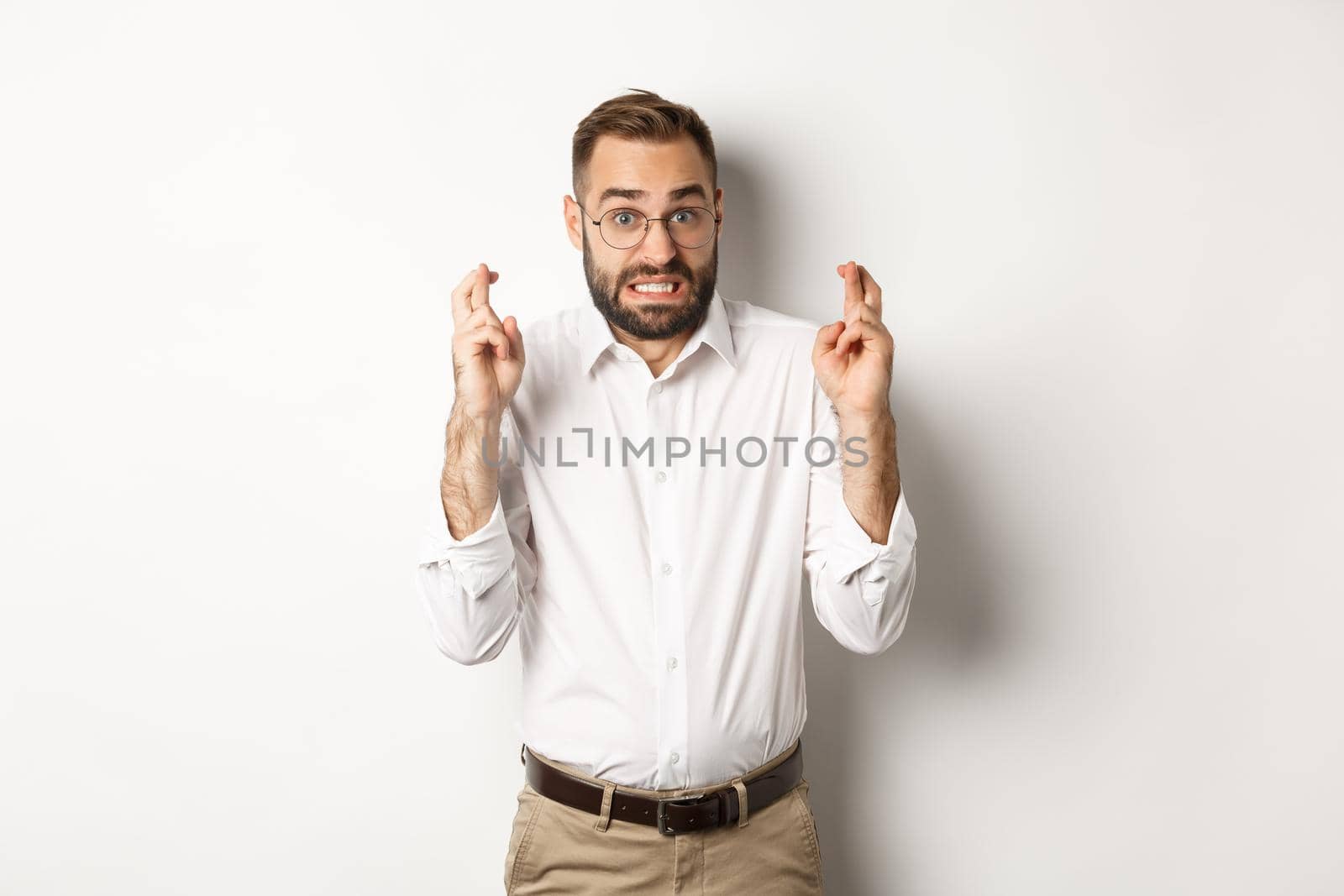 Worried man making a wish, cross fingers and hope for relish, standing over white background.