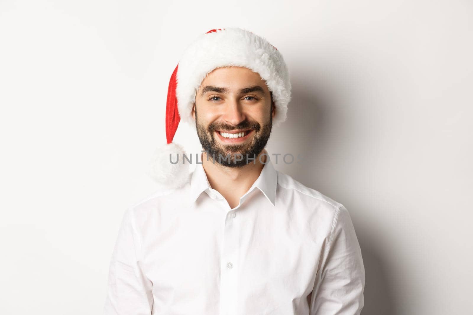 Close-up of happy bearded man celebrating christmas, wearing santa party hat and smiling, standing over white background by Benzoix