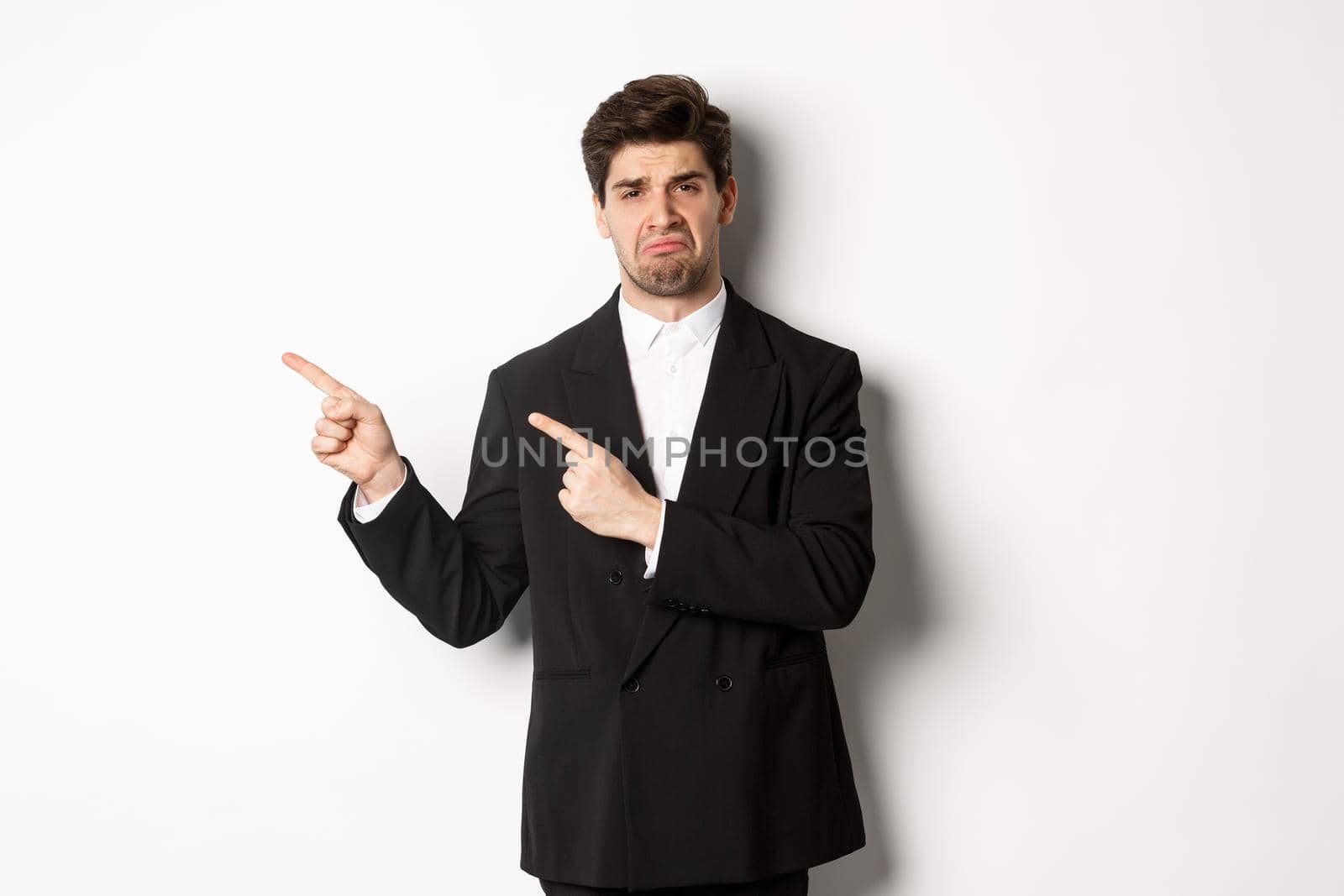 Portrait of disappointed and sad handsome businessman in suit, complaining and pointing fingers left at something bad, standing upset against white background.