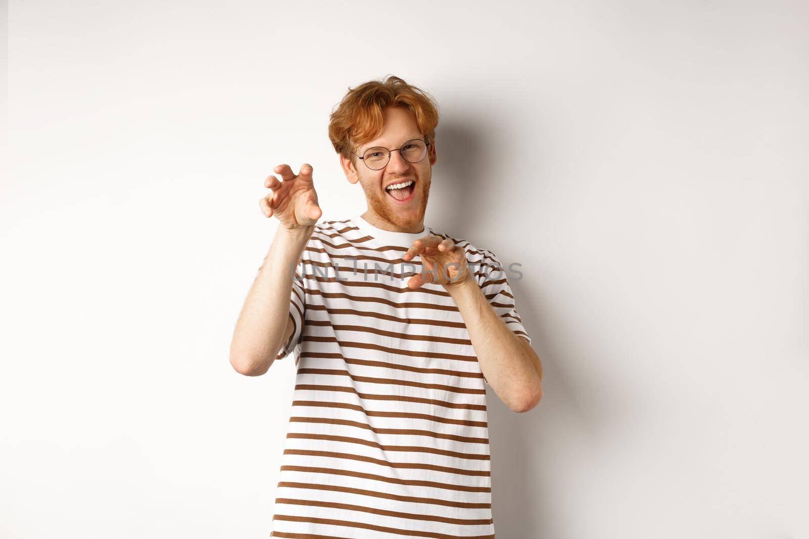 Funny young man with red hair and beard showing claws gesture and roaring, having fun, standing happy over white background by Benzoix