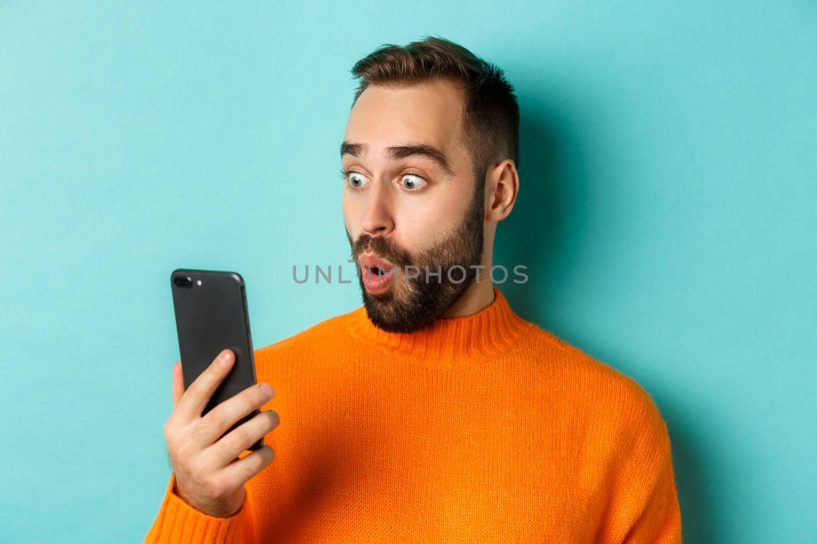 Close-up of caucasian man staring at phone screen with surprised face, wearing orange sweater, light blue background.
