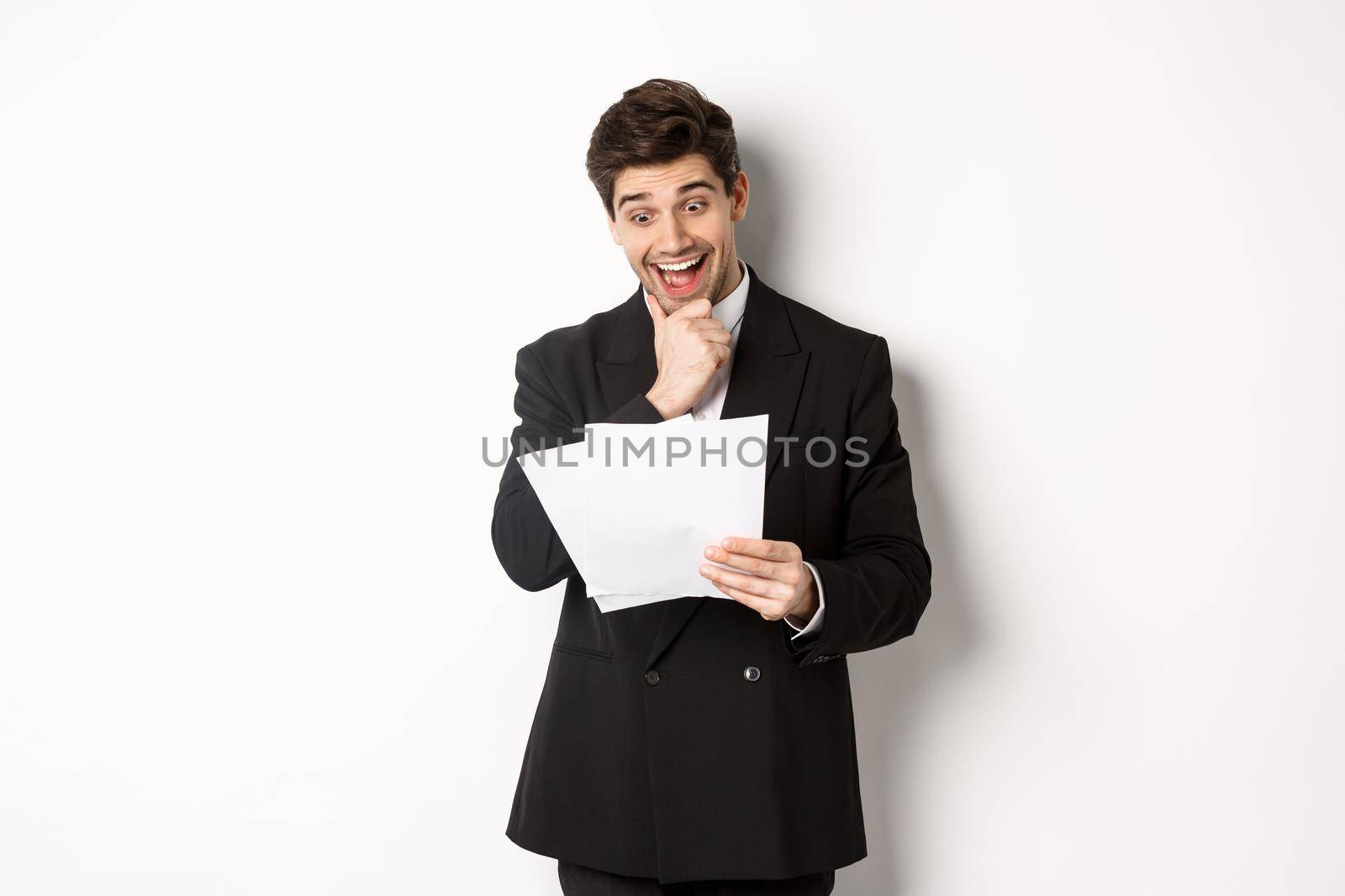 Portrait of handsome businessman looking excited at documents, working, standing against white background in black suit.