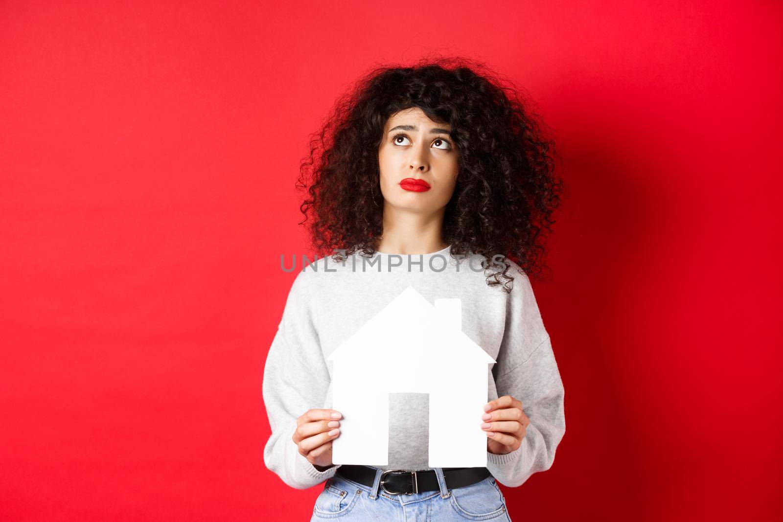 Real estate. Sad woman dreaming of buying apartment, holding paper house cutout and looking up distressed, standing on red background.