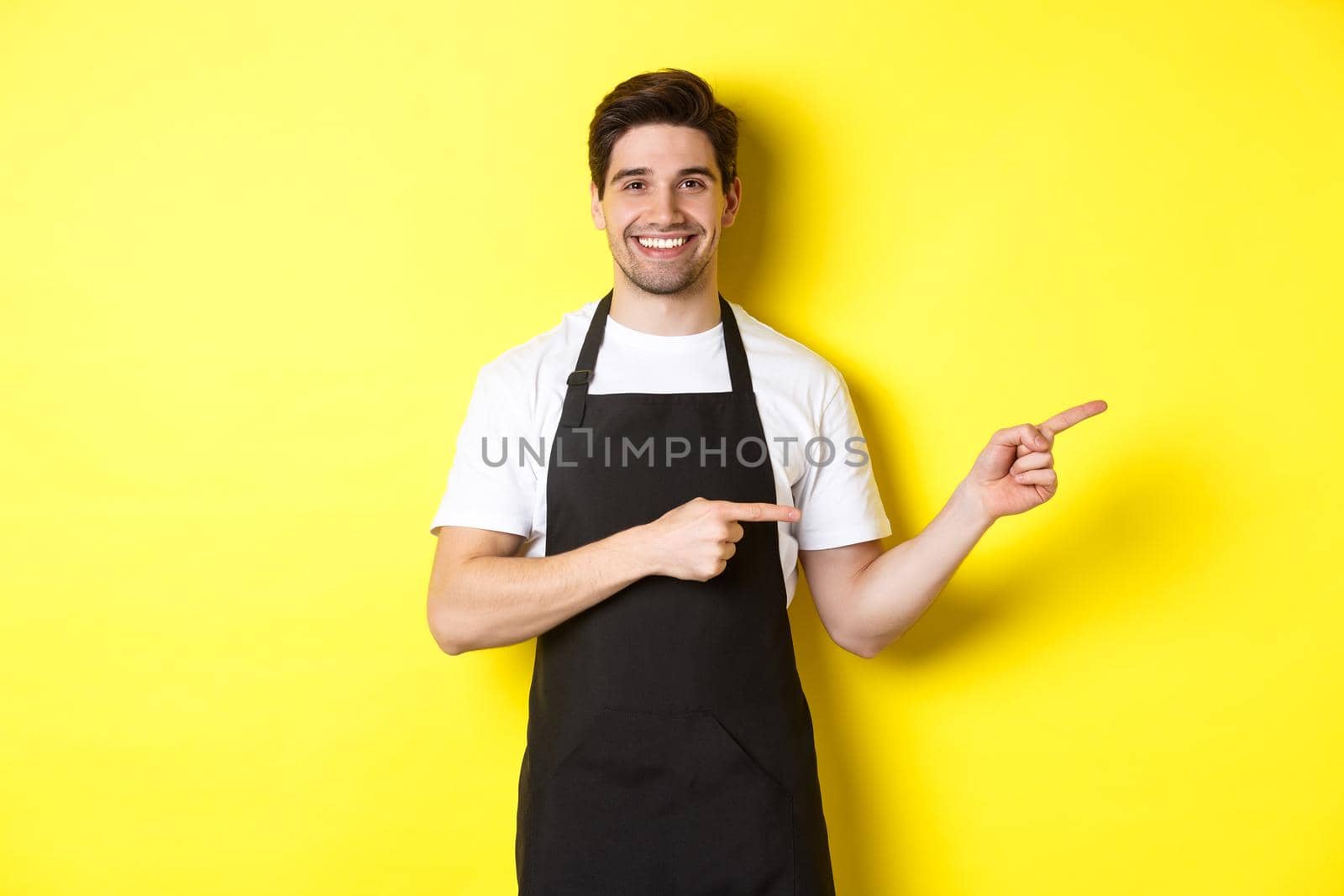 Friendly waiter pointing fingers right, showing your logo or promo offer, wearing black apron uniform, standing over yellow background by Benzoix