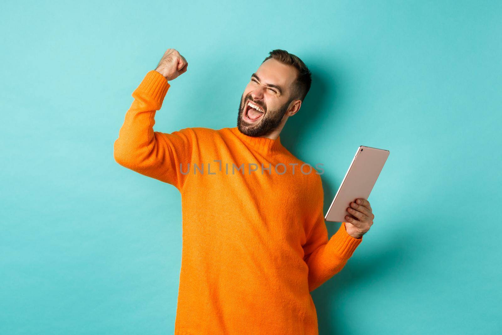 Cheerful winning man holding digital tablet, rejoicing and celebrating victory in game, making fist pump gesture, standing over light blue background.