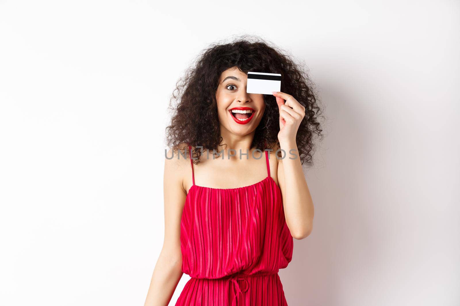 Shopping. Excited curly girl in red dress, showing plastic credit card and scream from joy, amazed with discounts and prices, standing against white background.