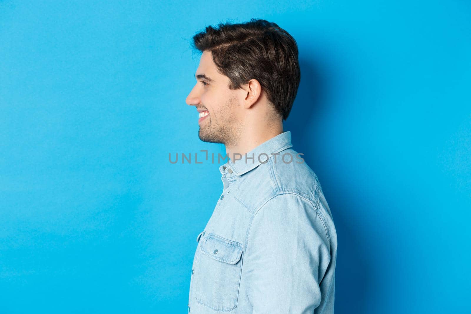 Profile of handsome young man looking left, smiling happy, standing over blue background.