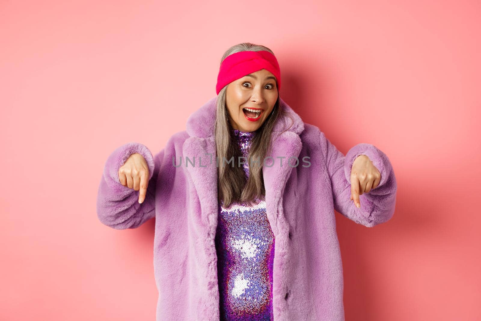 Fashion and shopping concept. Excited asian elderly woman checking out special deal, pointing fingers down and smiling happy at camera, showing advertisement, pink background by Benzoix