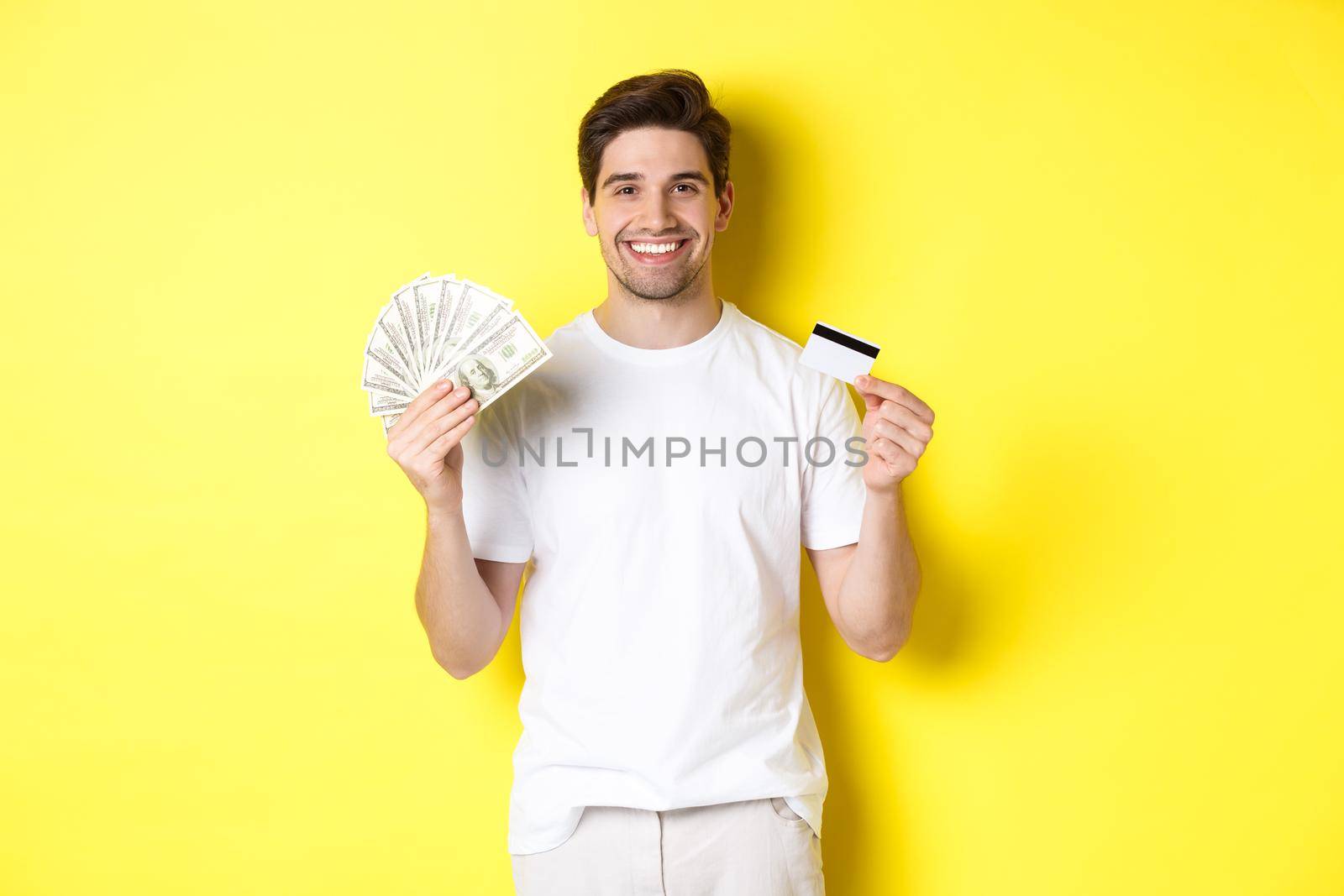 Young man withdraw money from credit card, smiling pleased, standing over yellow background.