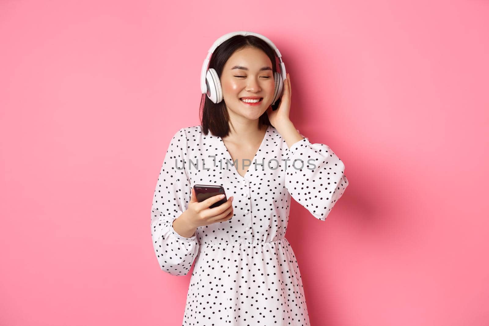 Romantic asian girl listening music in headphones, smiling with closed eyes, holding mobile phone, standing over pink background.