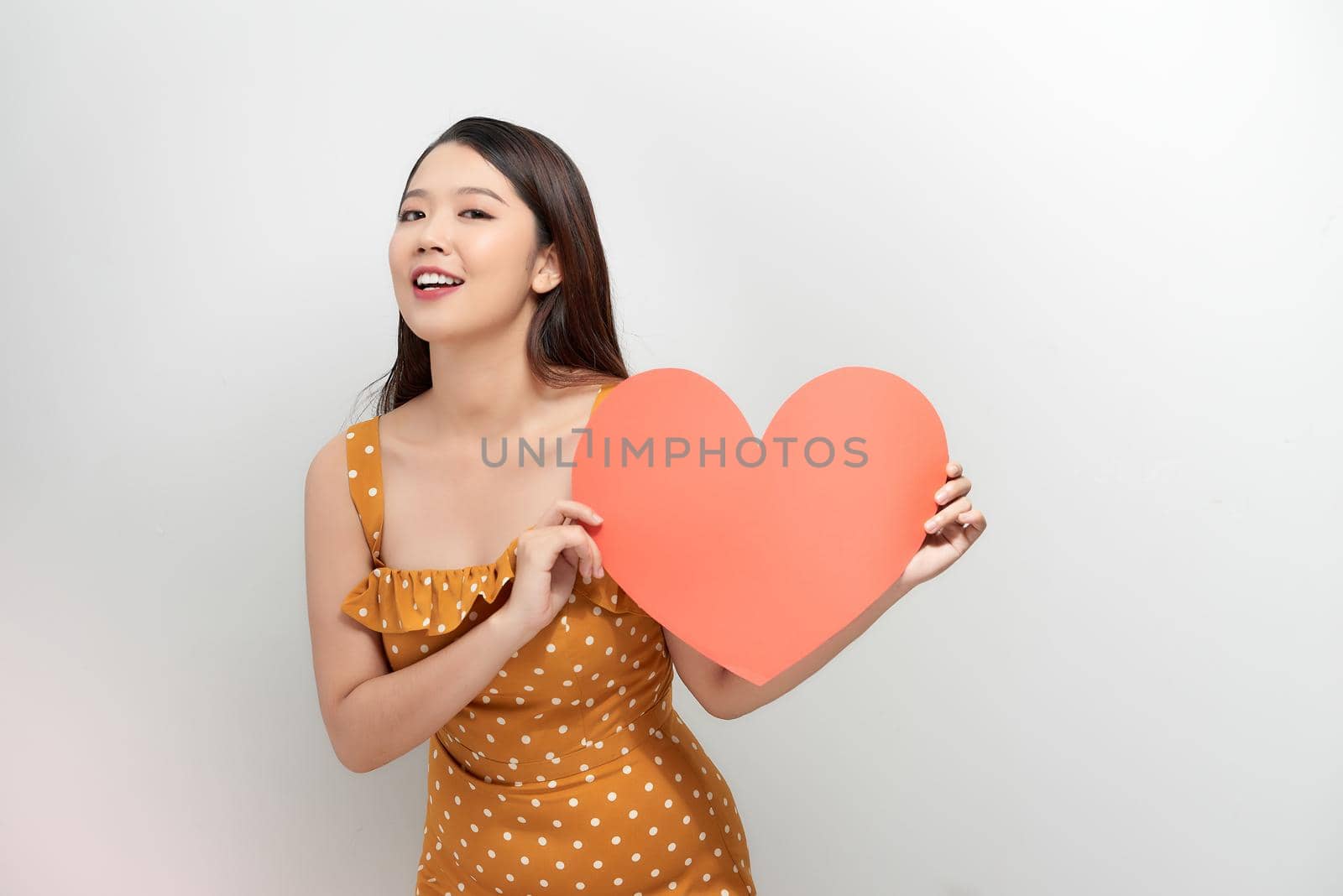 smiling pretty woman looking upwards, holding large red heart to chest daydreaming of men in love, by makidotvn