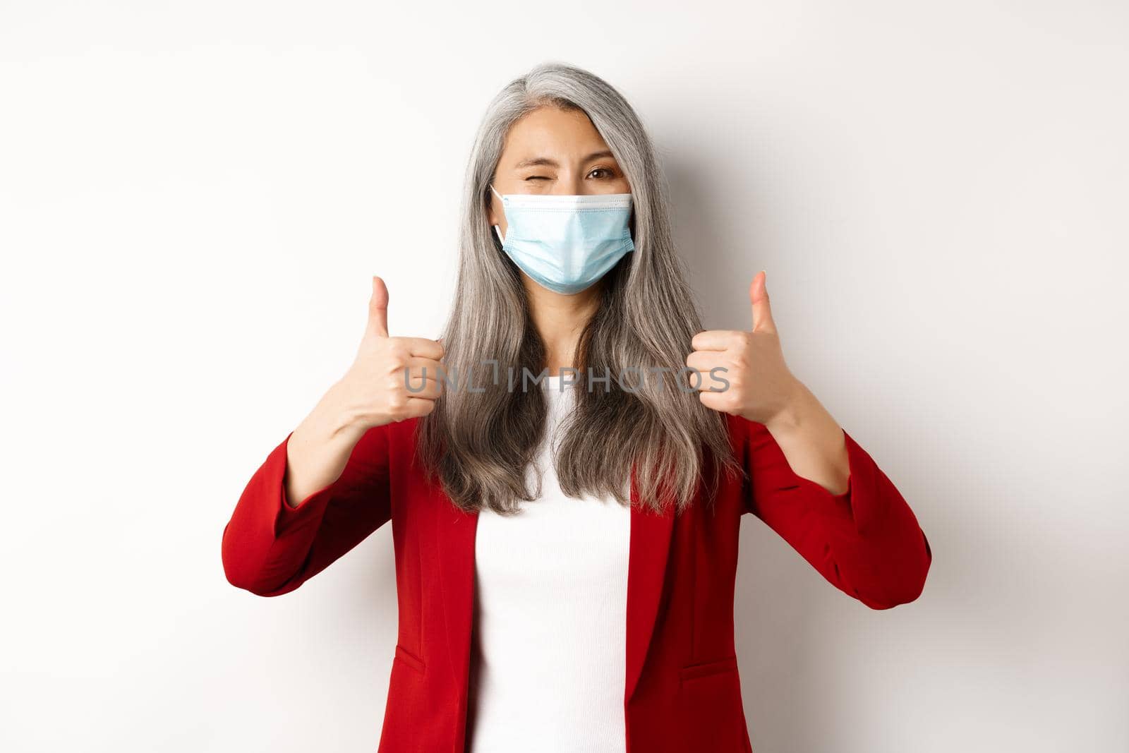 Coronavirus and business concept. Asian female entrepreneur in face mask looking cheerful, showing thumb-up in approval, white background.