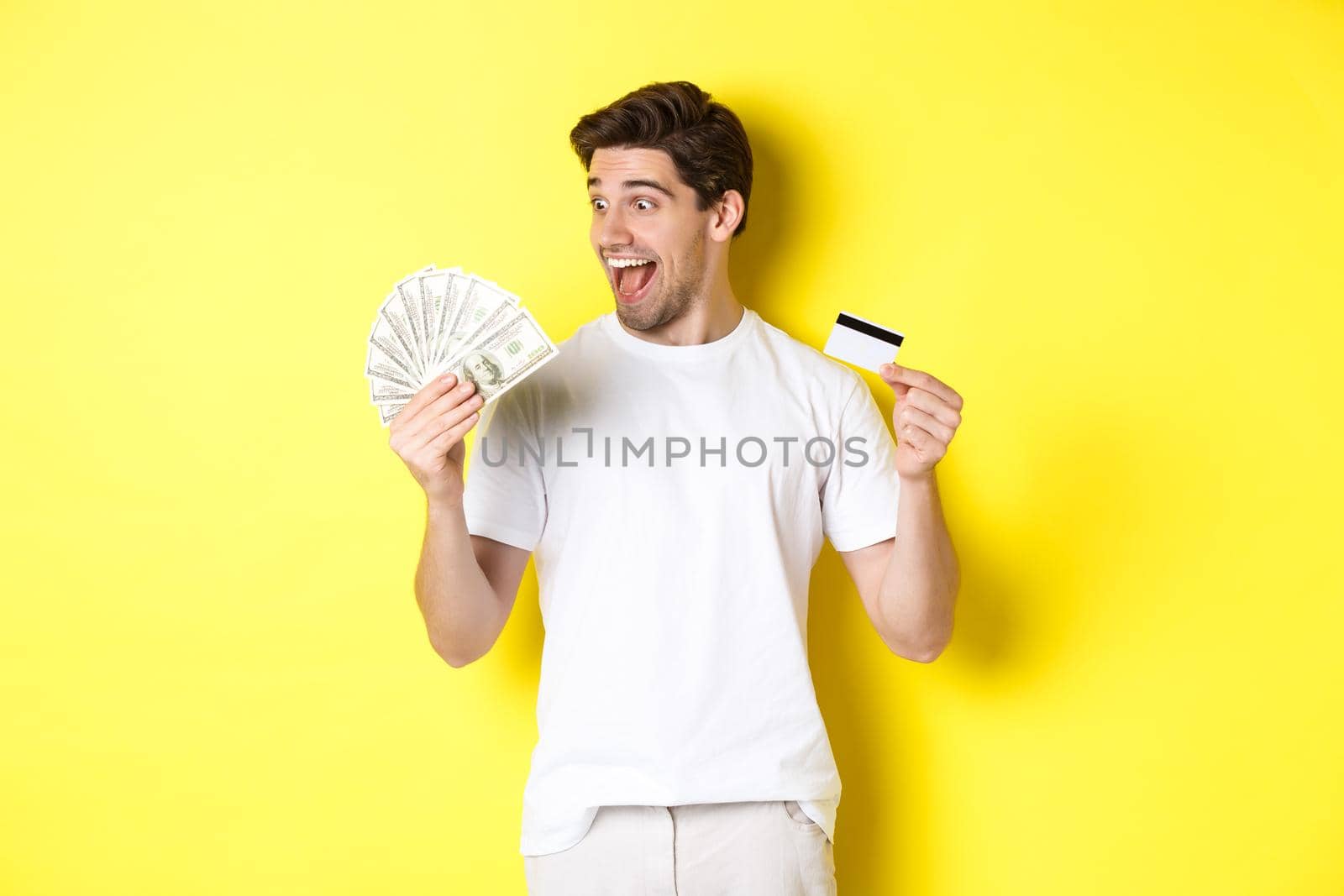Cheerful guy looking at money, holding credit card, concept of bank credit and loans, standing over yellow background by Benzoix