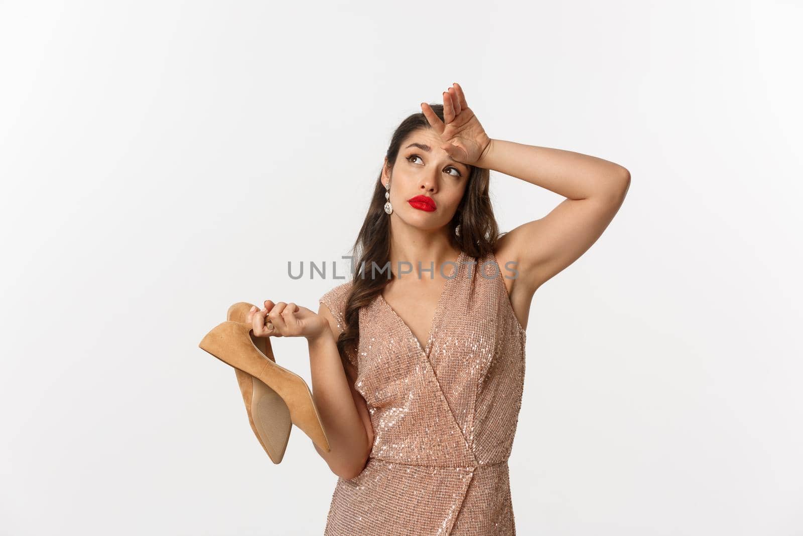 Party and celebration concept. Tired woman in elegant dress take off pair of heels, holding hand on forehead, express fatigue, standing over white background by Benzoix