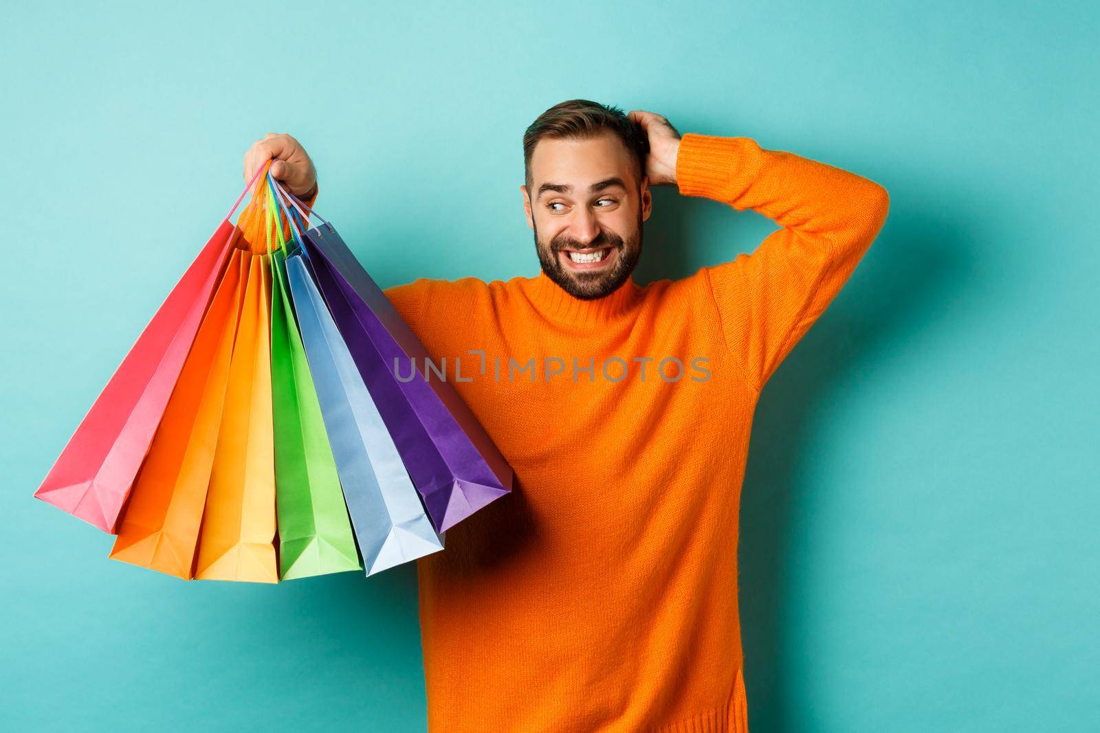 Handsome young man looking awkward and scratching head, staring at shopping bags with gifts, standing over turquoise background.