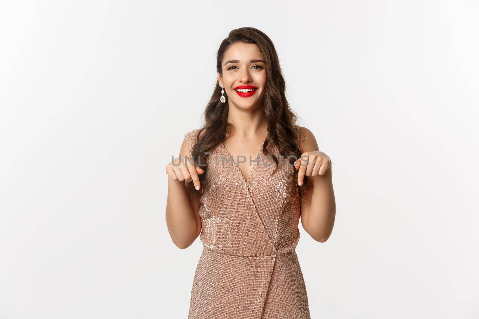 Happy beautiful woman with red lips, smiling while showing promo, wearing elegant dress for christmas party, pointing fingers down, standing over white background.